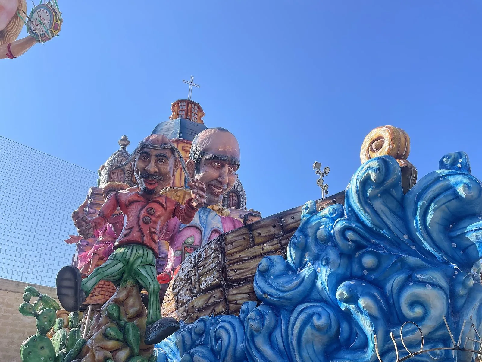 Carro allegorico in cartapesta con grandi caricature, onde blu, fichi d'India e una chiesa con cupola sotto il cielo azzurro