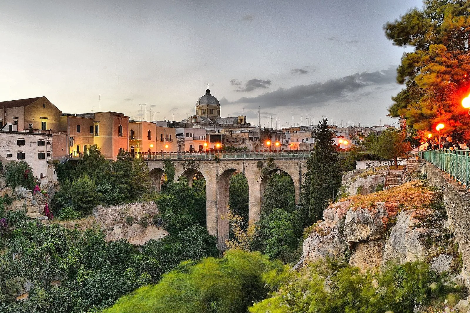 Ponte ad arcate in pietra sulla gravina verde, con il centro storico di Massafra e la cupola del duomo illuminati al tramonto